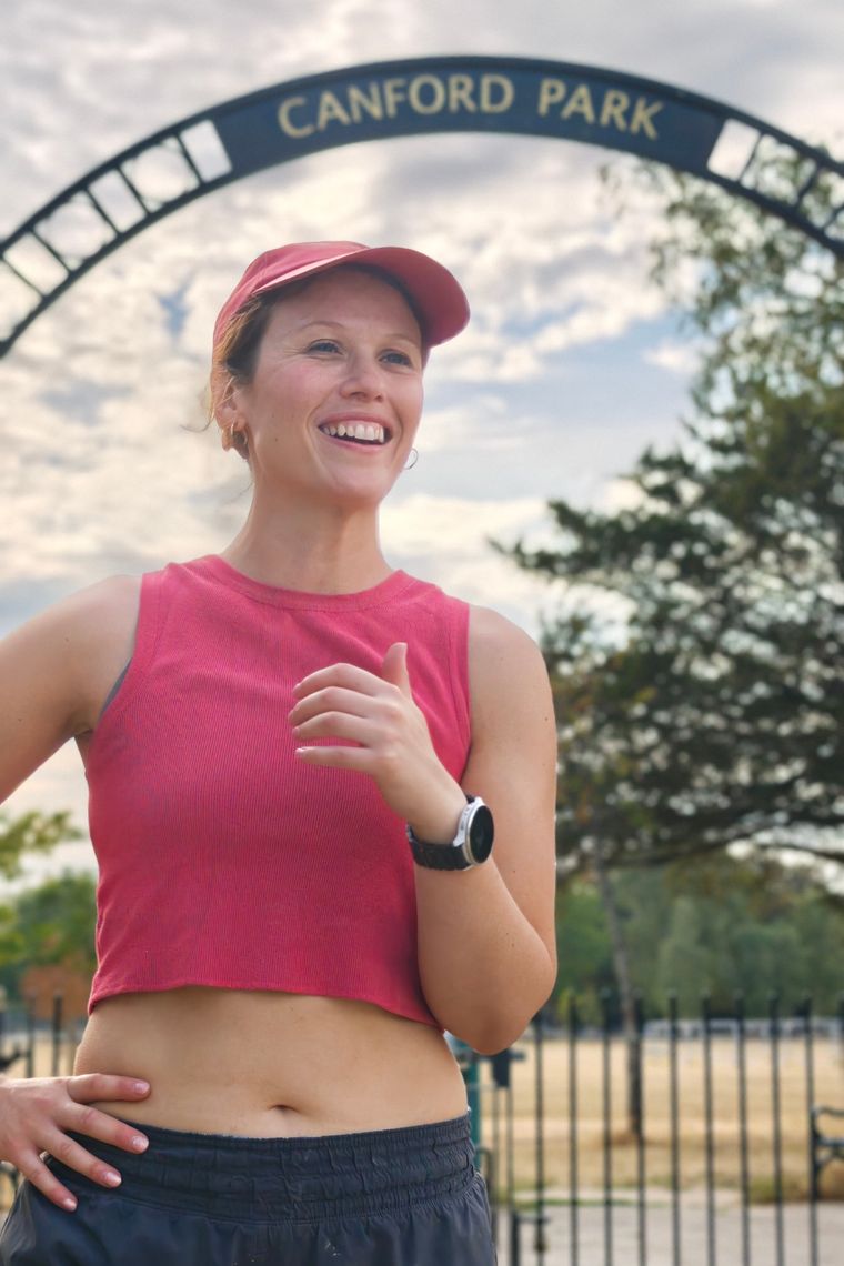Rose smiling in a pink vest and cap, standing under the wrought-iron Canford Park arch.